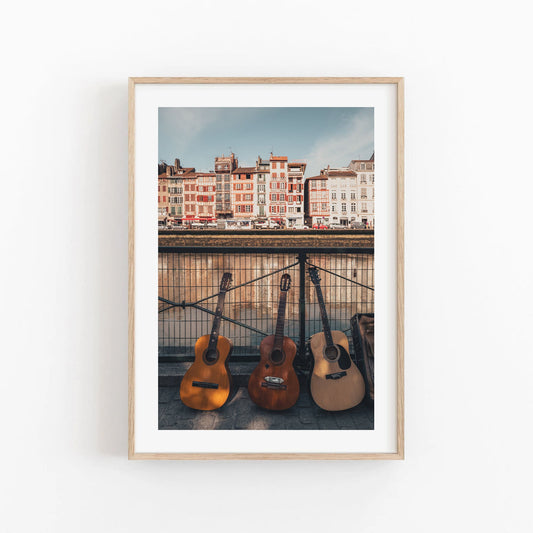 Framed photo of three guitars leaning against a waterfront railing with colorful buildings in the background.