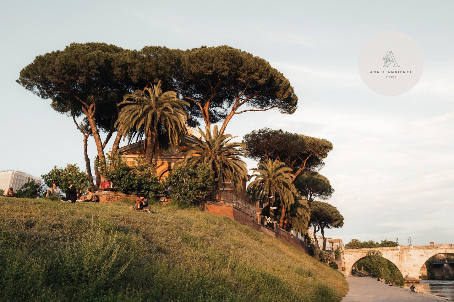 People enjoying Tiber Island Delight I on a grassy hill near trees and a stone bridge under a clear sky.