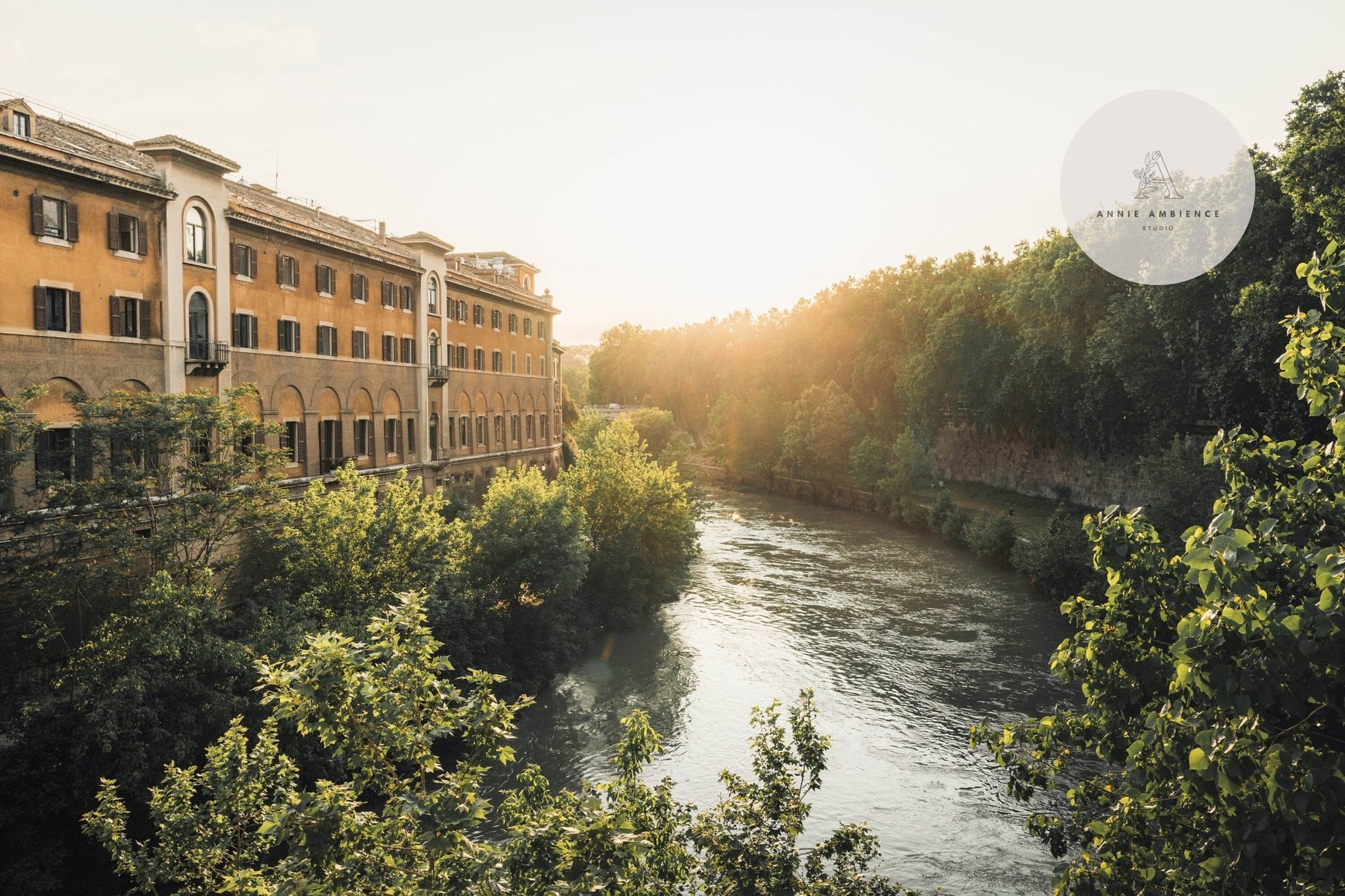 Experience the Tiber Island Glow II: a tranquil riverside view with an old building, lush greenery, and warm sunlight.