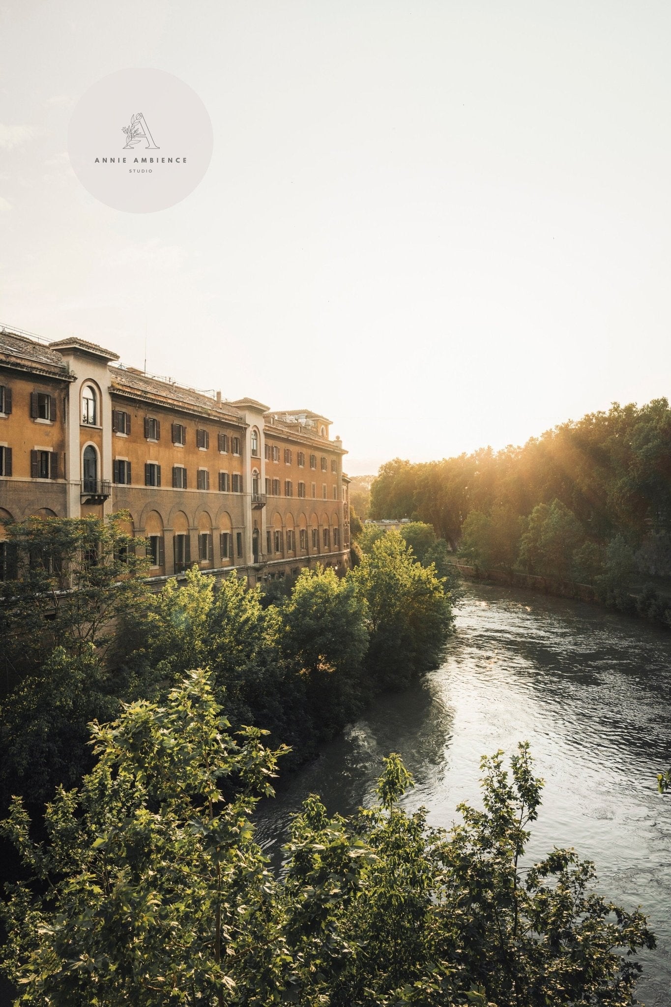 Tiber Island Glow: Riverside building with arches, trees, and river reflecting the warm glow of sunset.