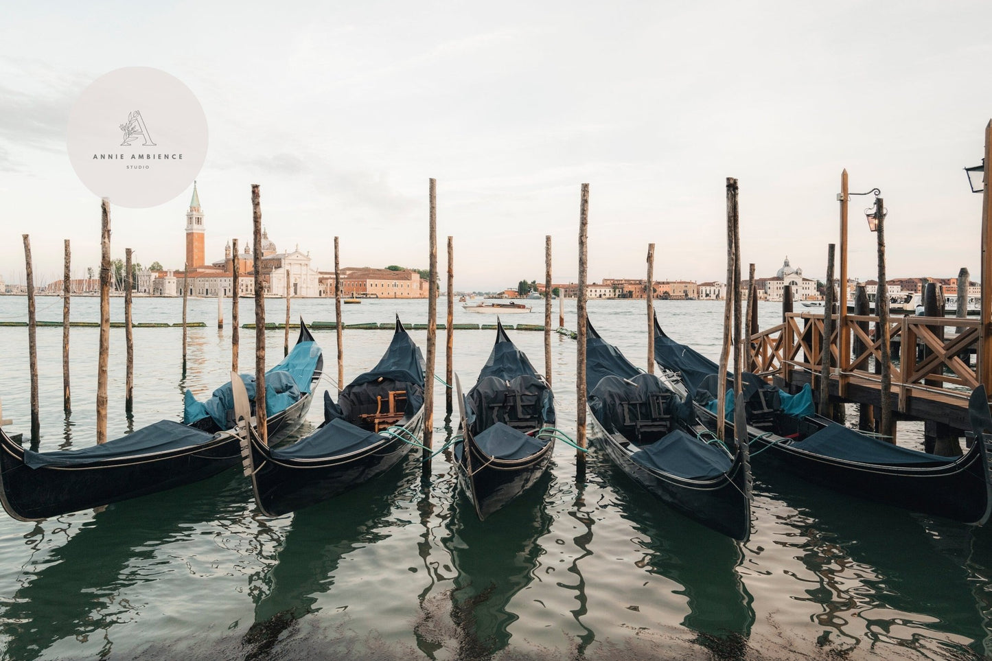 Five gondolas from the Venetian Array docked at a wooden pier, overlooking calm waters and historic buildings.