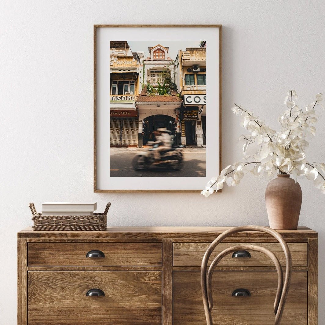 Pace of Hanoi framed photo hangs above a wooden dresser with white flowers in a vase and a wicker basket.