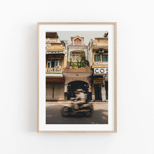 Framed photo titled Pace of Hanoi captures a blurred motorbike passing old shops and balcony-lined buildings.