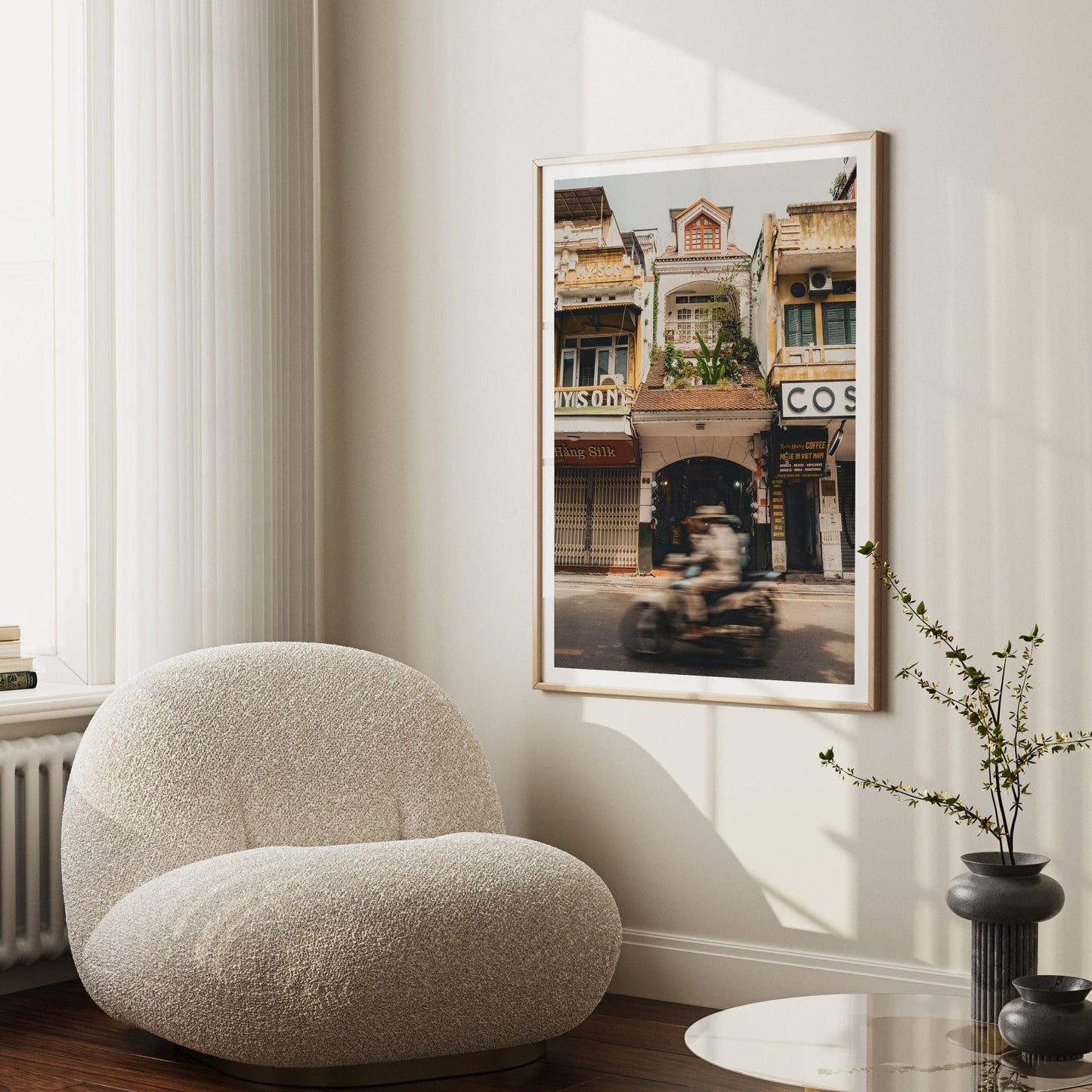 A cozy white chair sits by a window and table, with the Pace of Hanoi city street artwork on the wall.