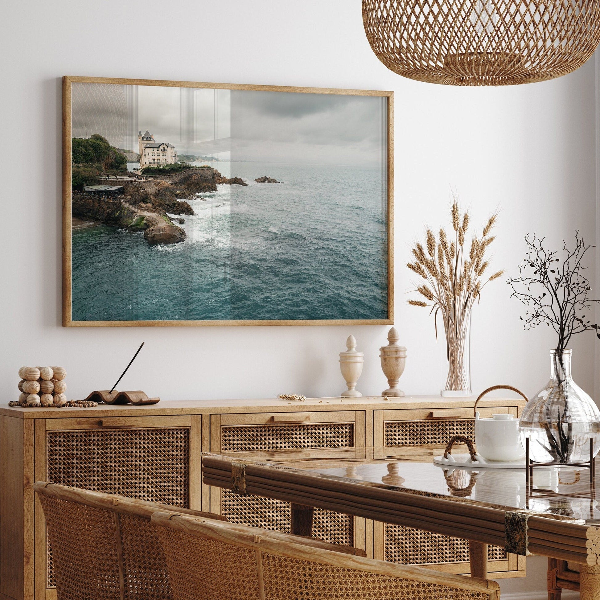 The Villa Beltza photo hangs in a dining room with wicker furniture, decorative jars, and a glass vase on the table.
