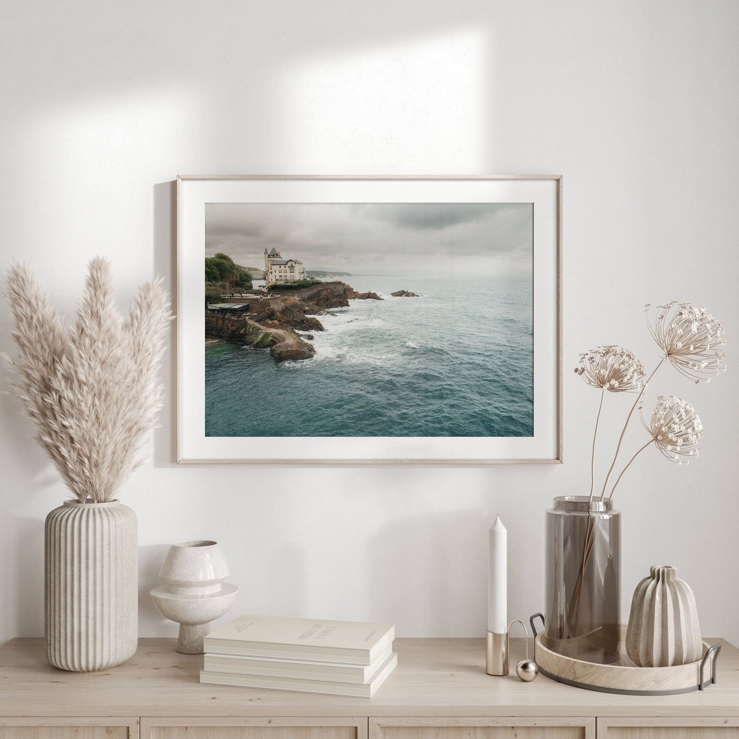 The Villa Beltza photo hangs above a wooden shelf with vases, dried plants, and a candle in a minimalistic room.