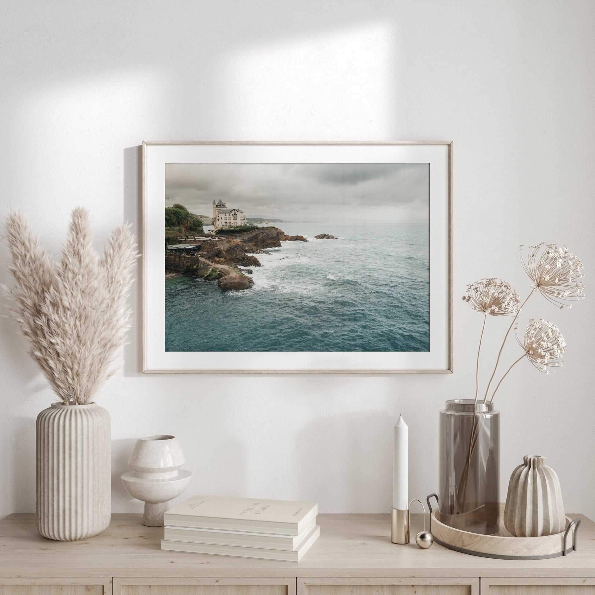 The Villa Beltza photo hangs above a wooden shelf with vases, dried plants, and a candle in a minimalistic room.