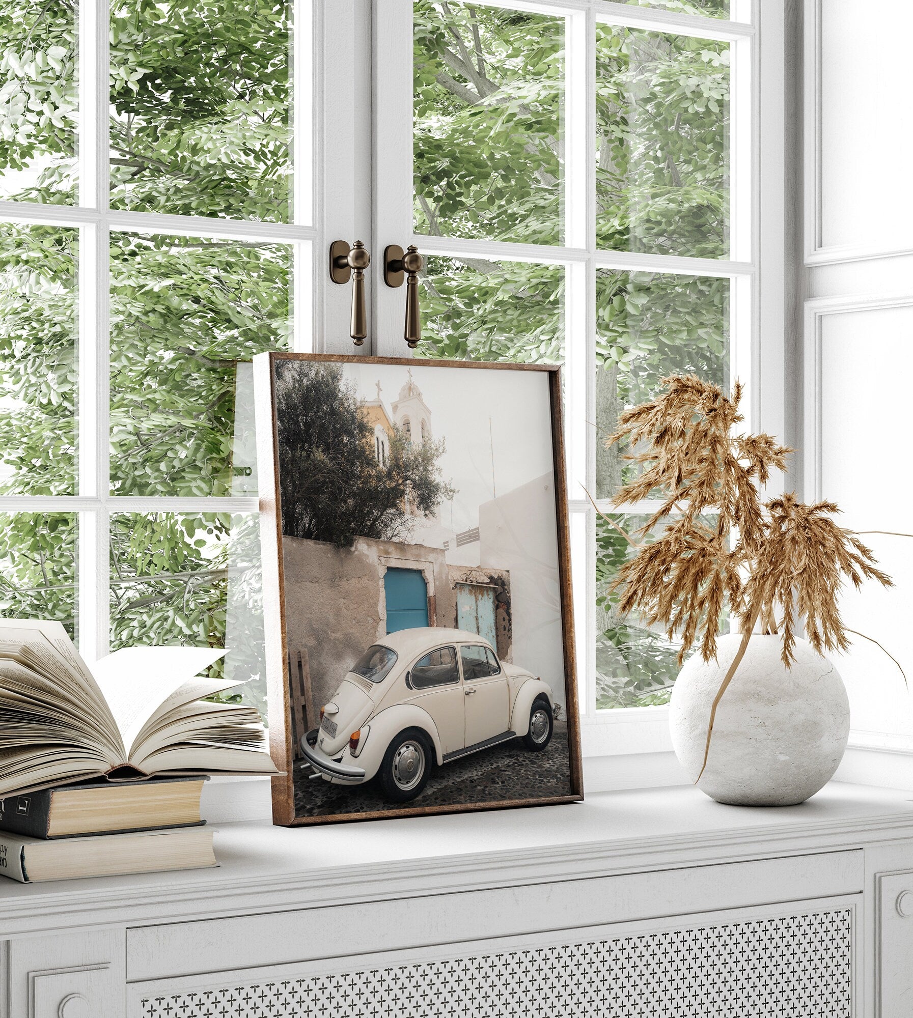 Framed photo of a Volkswagen Beetle and house on a windowsill with books, dried plants, and green foliage outside.