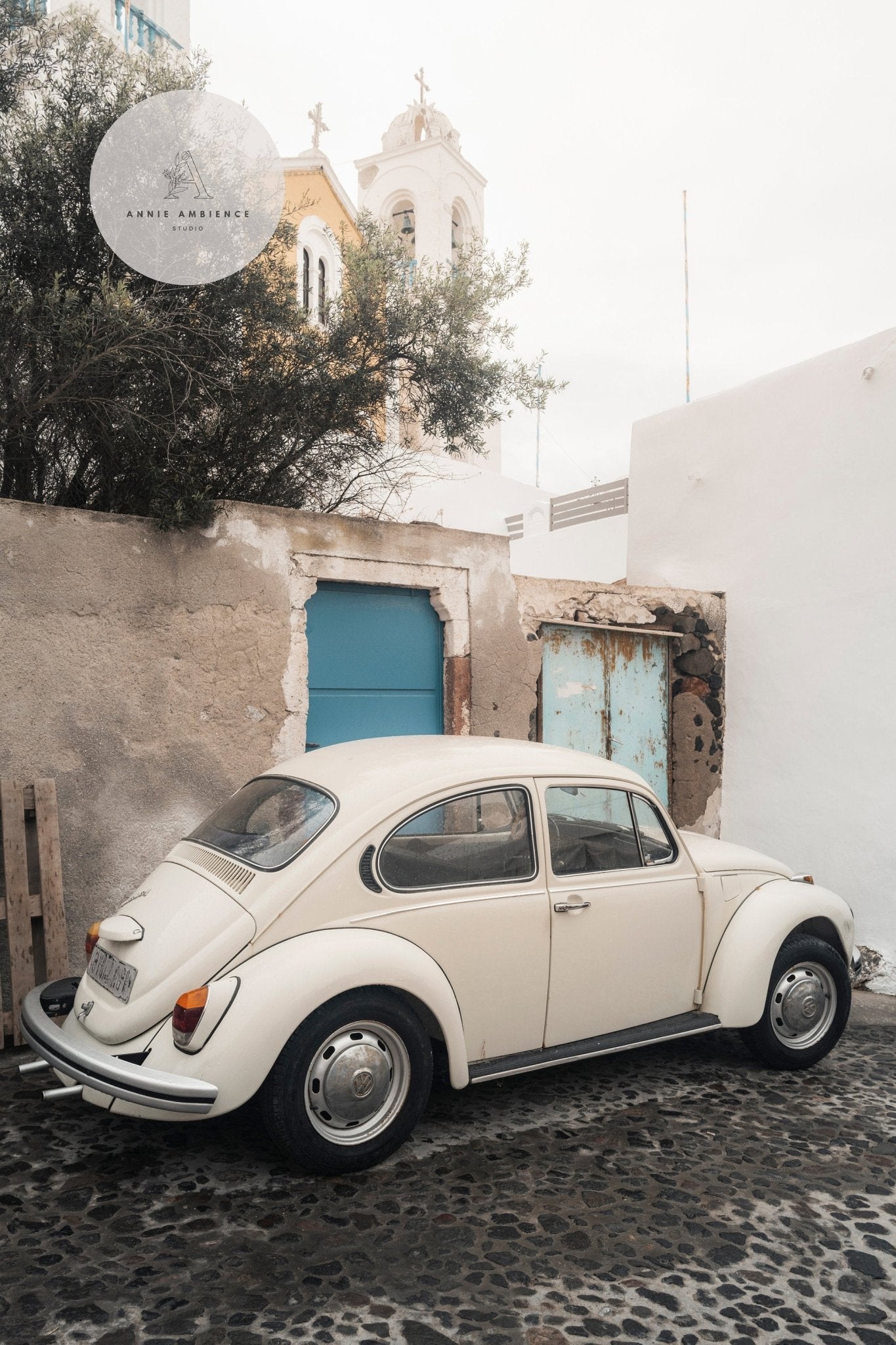 White Volkswagen Beetle beside a weathered wall and blue gate with a church tower in the background.