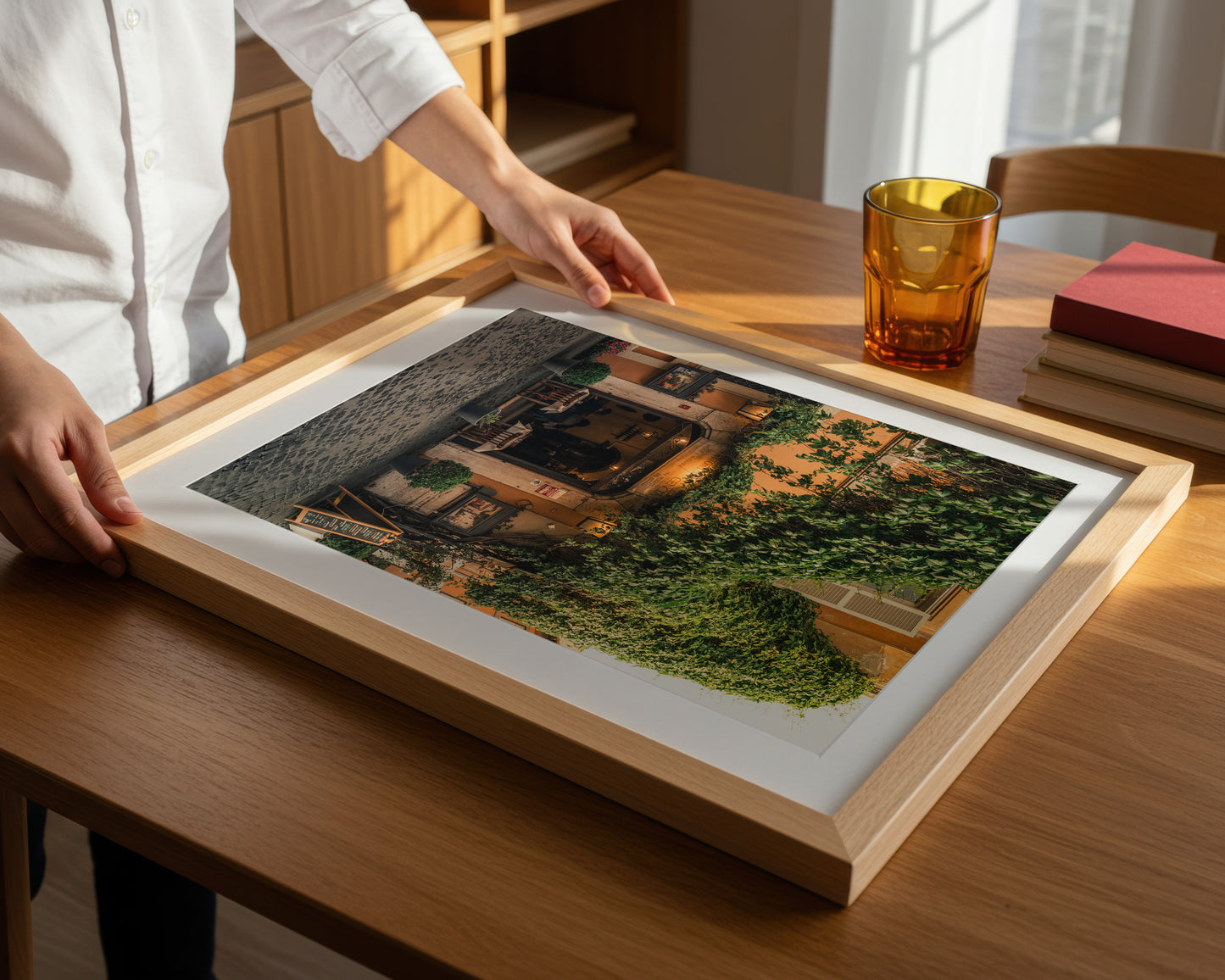 A person sets the Green Vines Trastevere framed photo on a wooden table next to a glass and books.