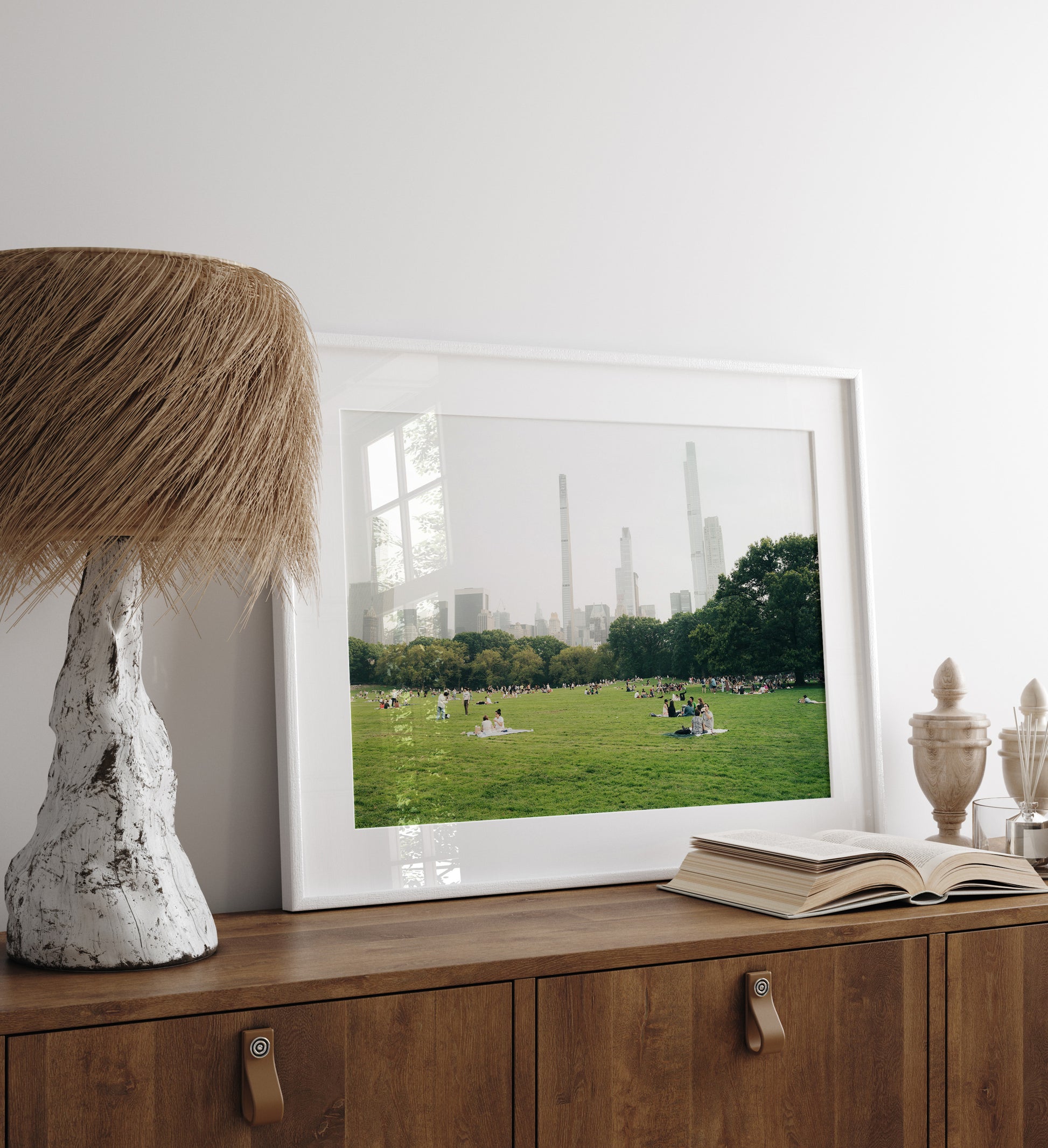 A framed "Great Lawn" city park photo, lamp, and book sit on a wooden cabinet against a white wall.