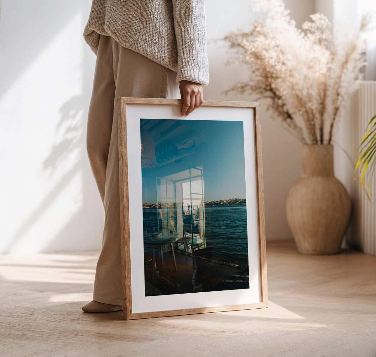 Person holding the “Almada Reflections” framed photo indoors near a vase with dried pampas grass.