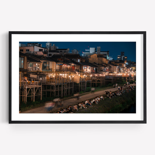 Framed photo of Kamo River at night, featuring illuminated buildings and people gathered along the riverbank.