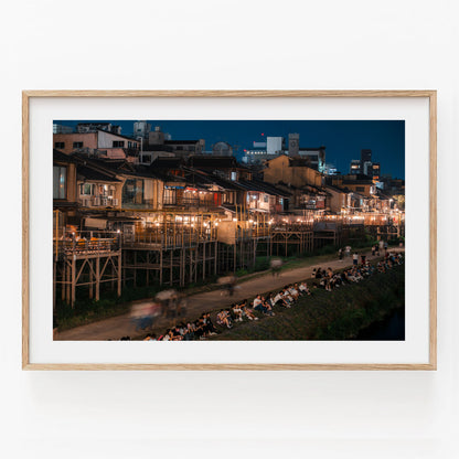 Kamo River framed nighttime cityscape with riverbank seating and lit buildings, displayed on a white wall.