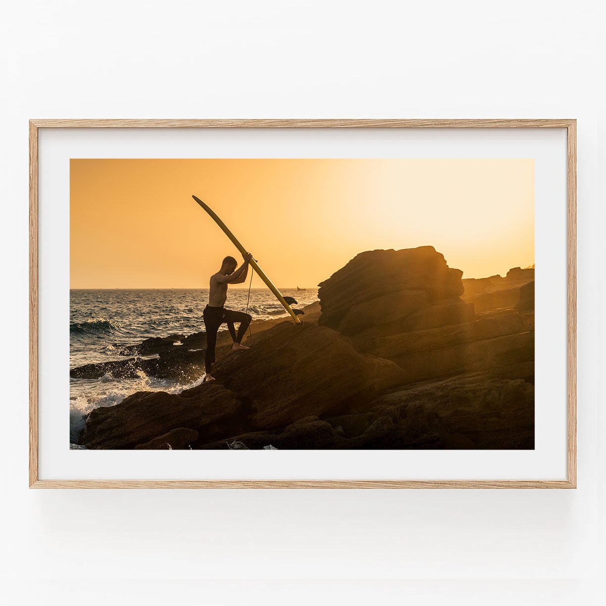 The Sunset Surfer stands on a rocky shore at sunset, surfboard in hand, as waves crash behind.