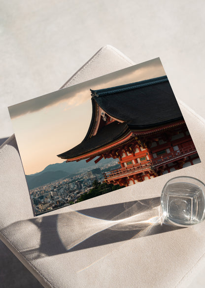 Kiyomizu-dera Sunset III print features a temple roof, cityscape, and mountains, displayed near a glass on cushion.