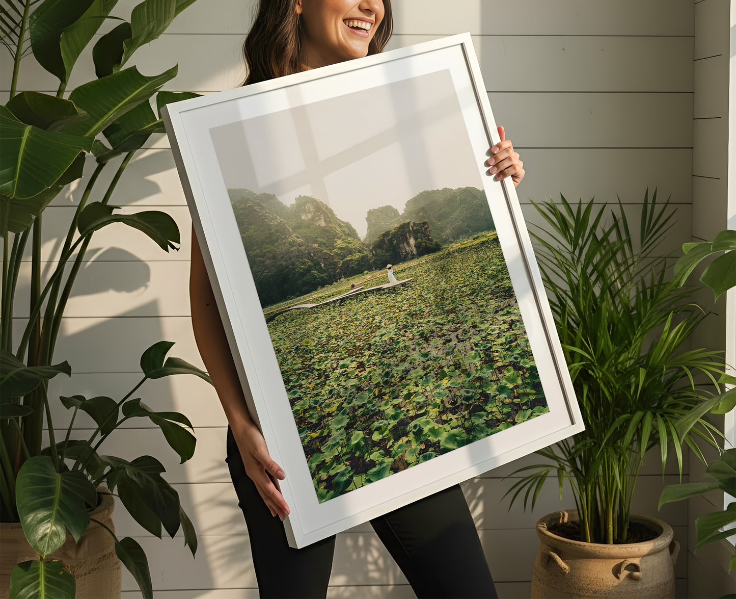 Woman smiles holding "Lotus Lady" framed photo of a lily pond, surrounded by indoor green potted plants.