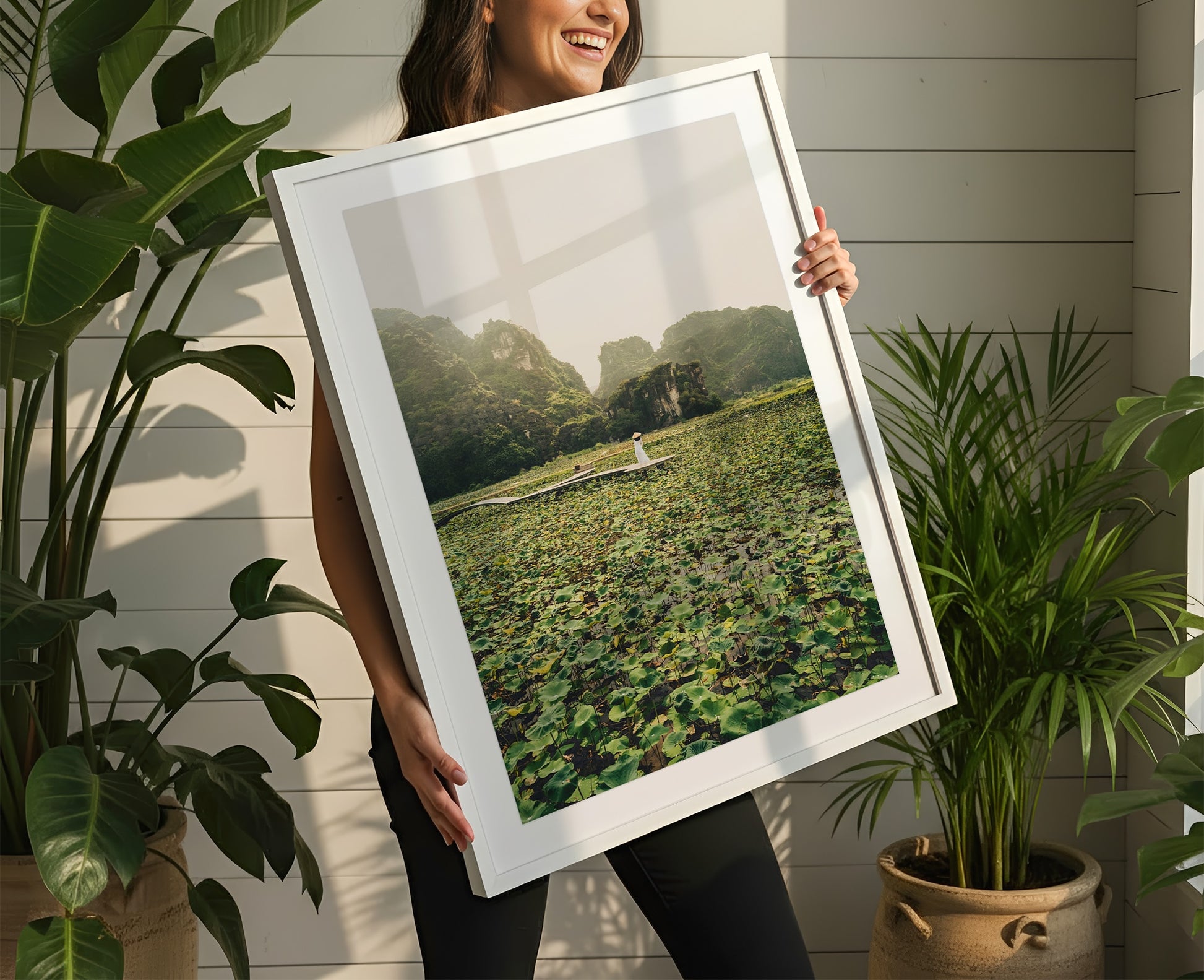 Woman smiles holding "Lotus Lady" framed photo of a lily pond, surrounded by indoor green potted plants.