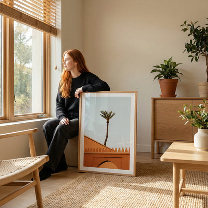 A woman in black sits by a window, holding a framed "Marrakesh Palm Tree" art print.