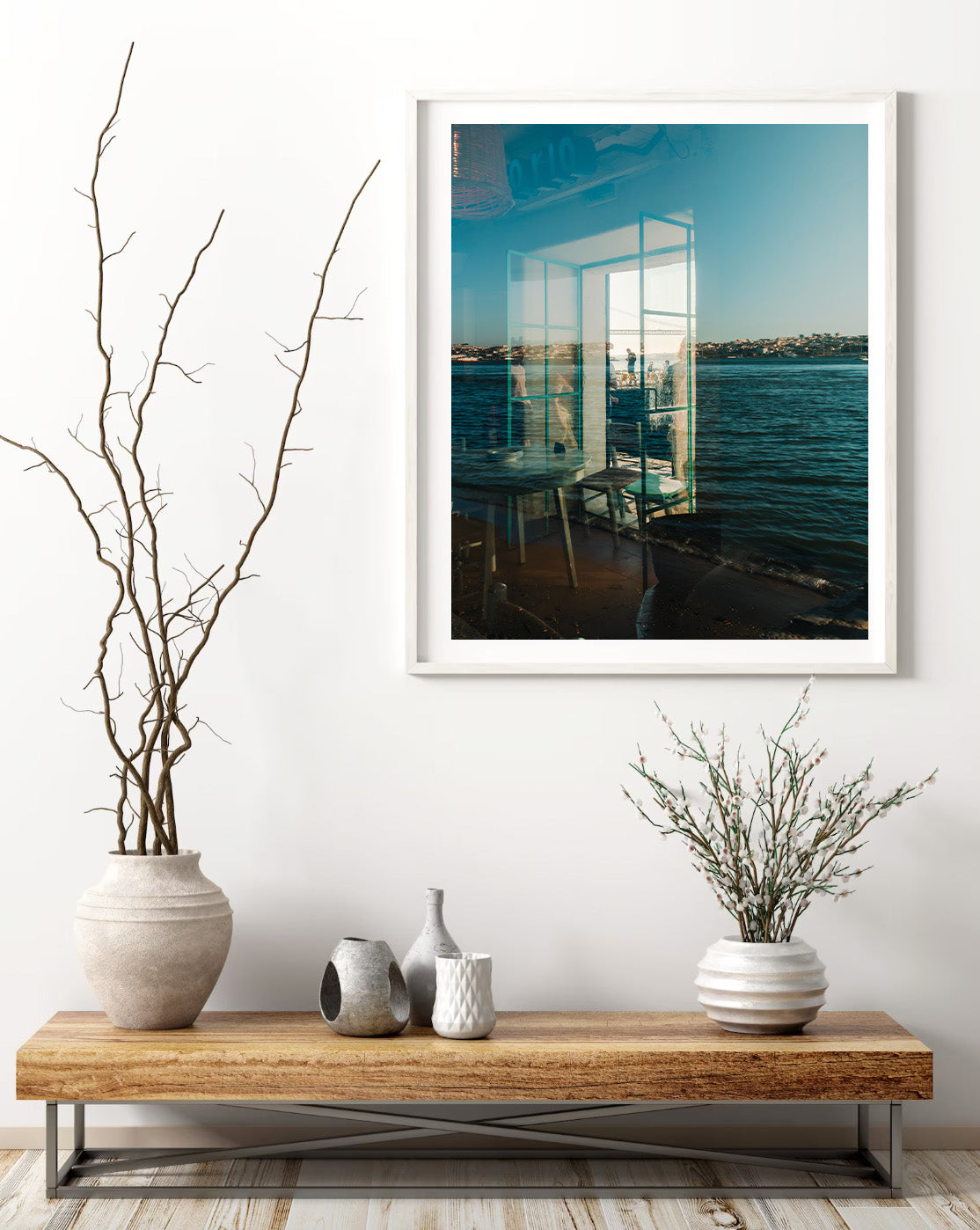 Almada Reflections framed photo of water and buildings above a table with vases, dried branches, and a white wall.