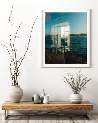 Almada Reflections framed photo of water and buildings above a table with vases, dried branches, and a white wall.