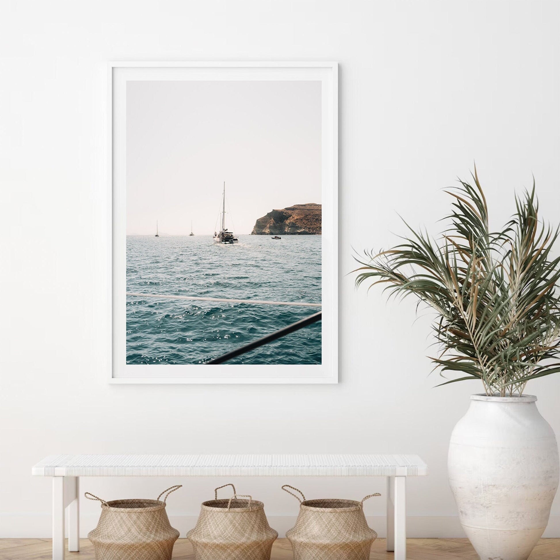 Afternoon Sail I framed sea view with sailboats on a white wall, above a bench, 3 woven baskets, and a large potted plant.