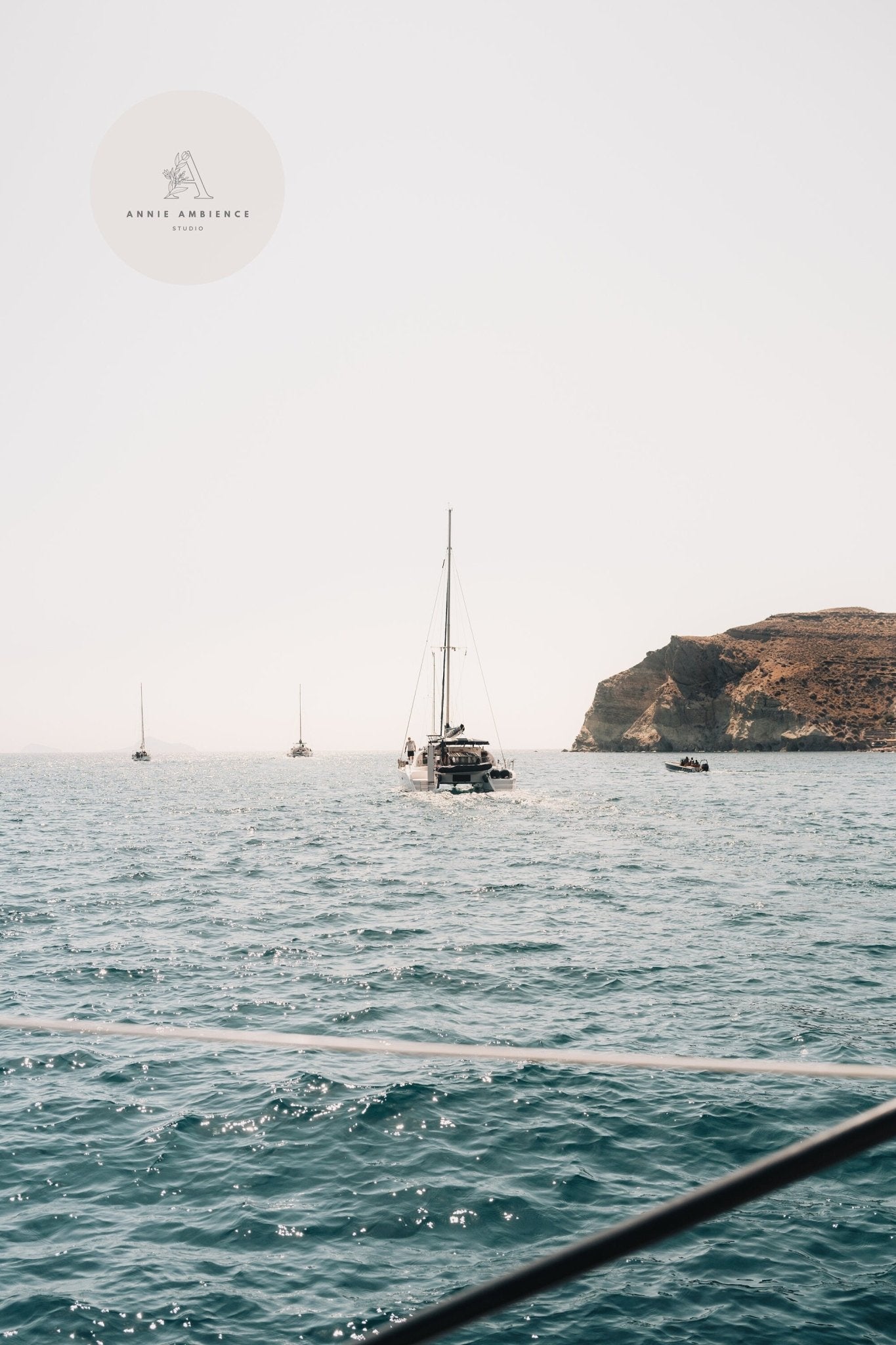 Afternoon Sail I: Sailboats glide on a calm sea, rocky coastline backdrop under a clear sky.