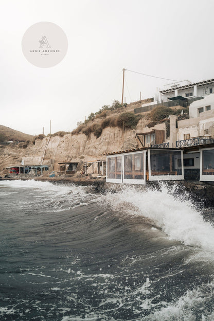 Akrotiri Wave: Rough waves crash against a rocky shoreline with buildings perched on a cliff.