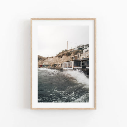 Framed Akrotiri Wave photo: waves crash against a rocky shore with cliffside buildings.