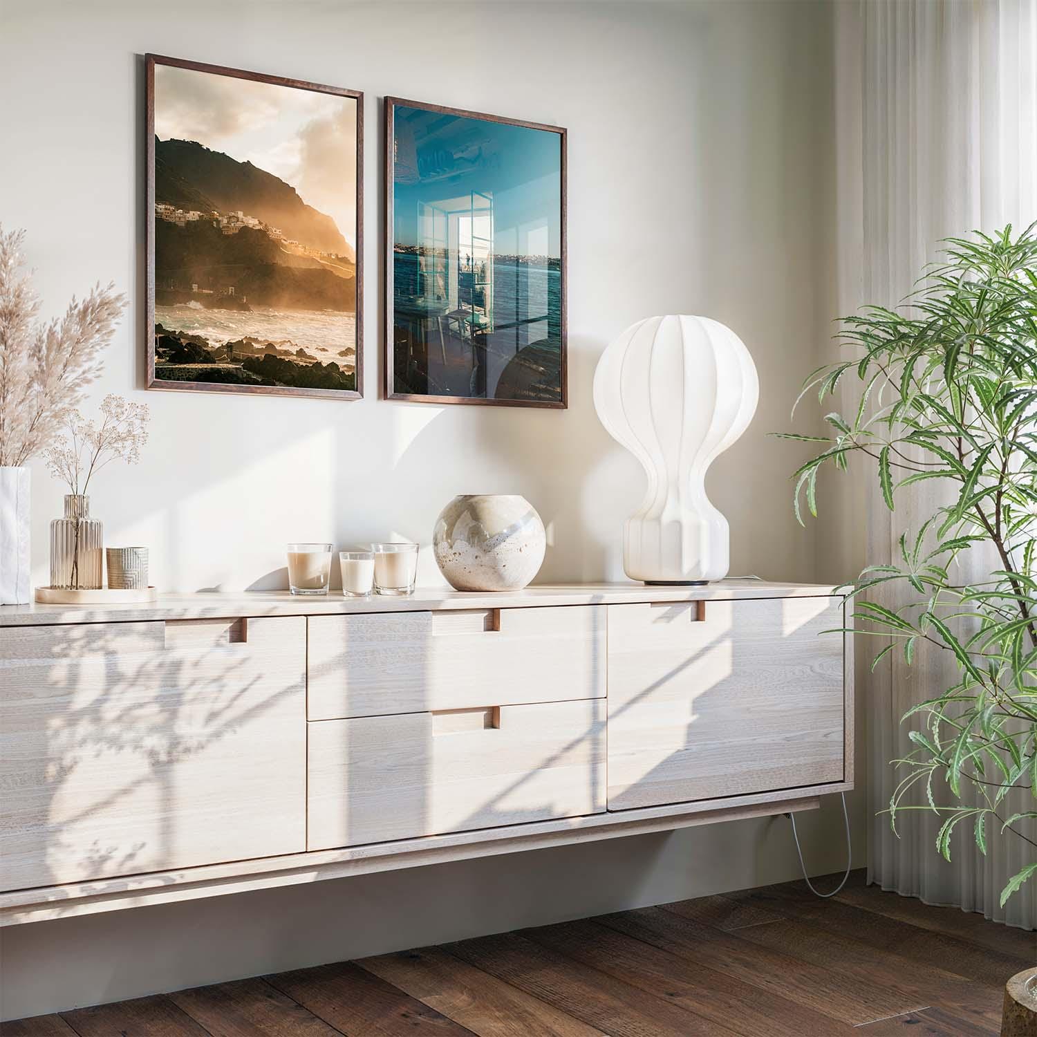 Modern sideboard with decor, large plant, lamp, and Atmospheric Coasts - Set of 2 framed on the sunlit wall.