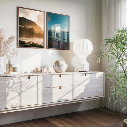 Modern sideboard with decor, large plant, lamp, and Atmospheric Coasts - Set of 2 framed on the sunlit wall.
