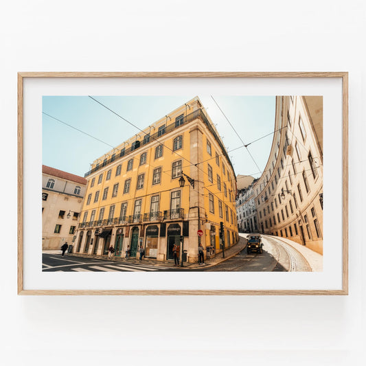 Framed photo of Baixa-Chiado: yellow building, curved street, tram, and people in a sunny European city.