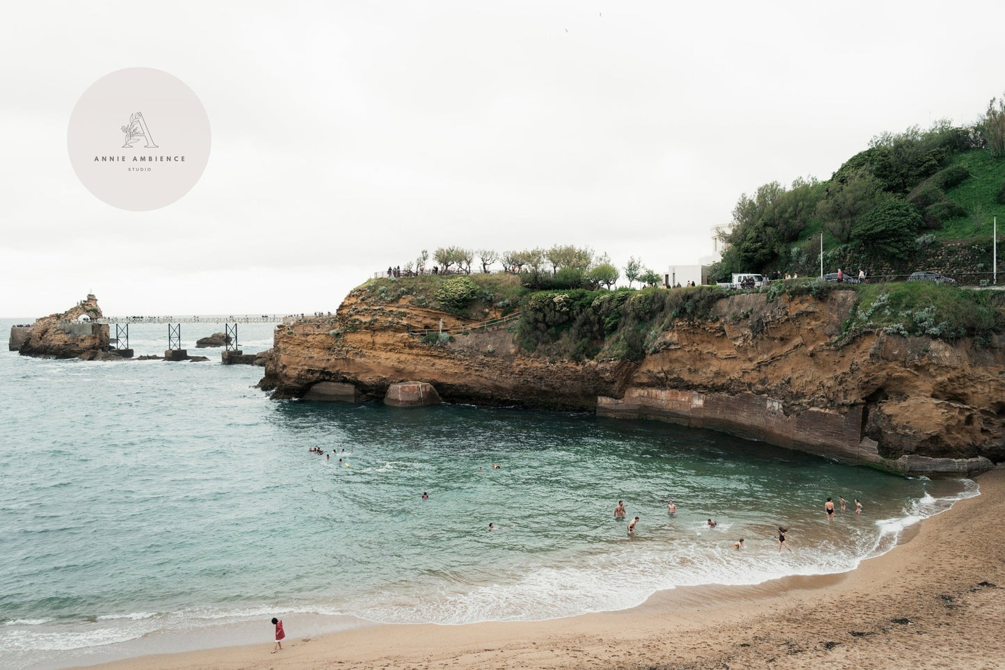 Biarritz Beach features swimmers near rocky cliffs under a cloudy sky, surrounded by greenery.