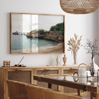 The Biarritz Beach painting is above a wooden sideboard in a stylish room with wicker furniture and dried plants.