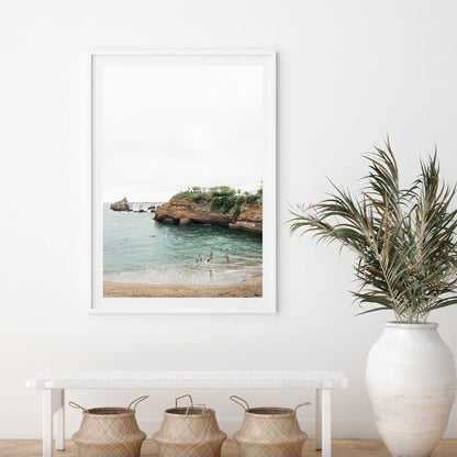 Biarritz Beach II framed photo on a white wall above a bench with baskets; potted plant next to the bench.