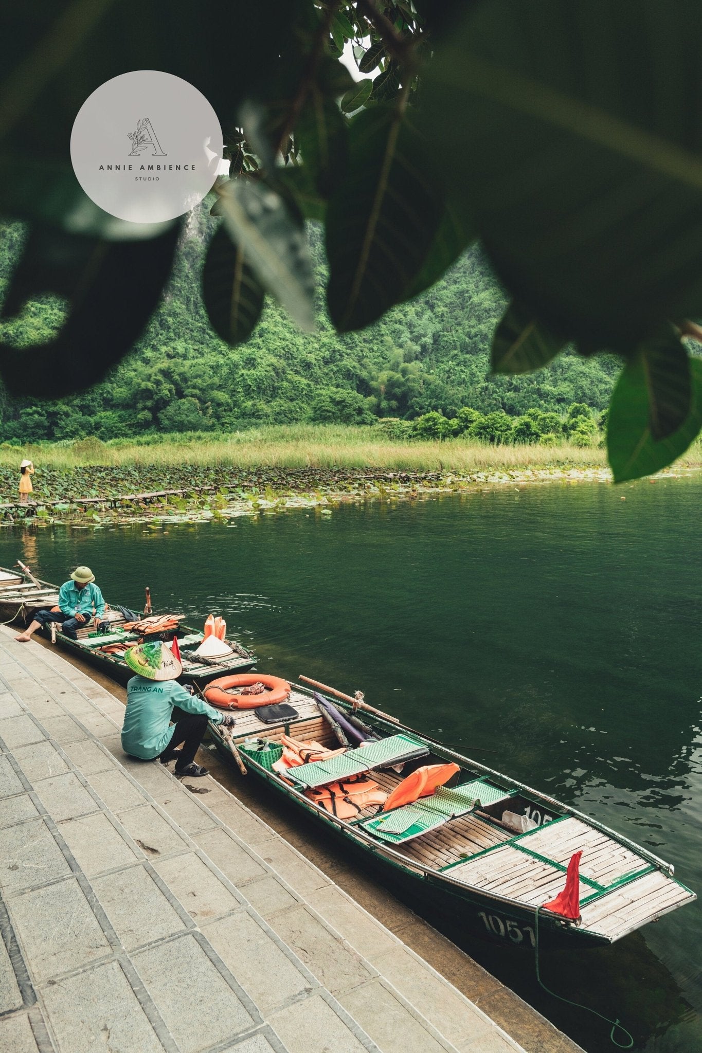 Two people in green hats enjoy Break Time on boats by a lush riverside, under overhanging leaves.