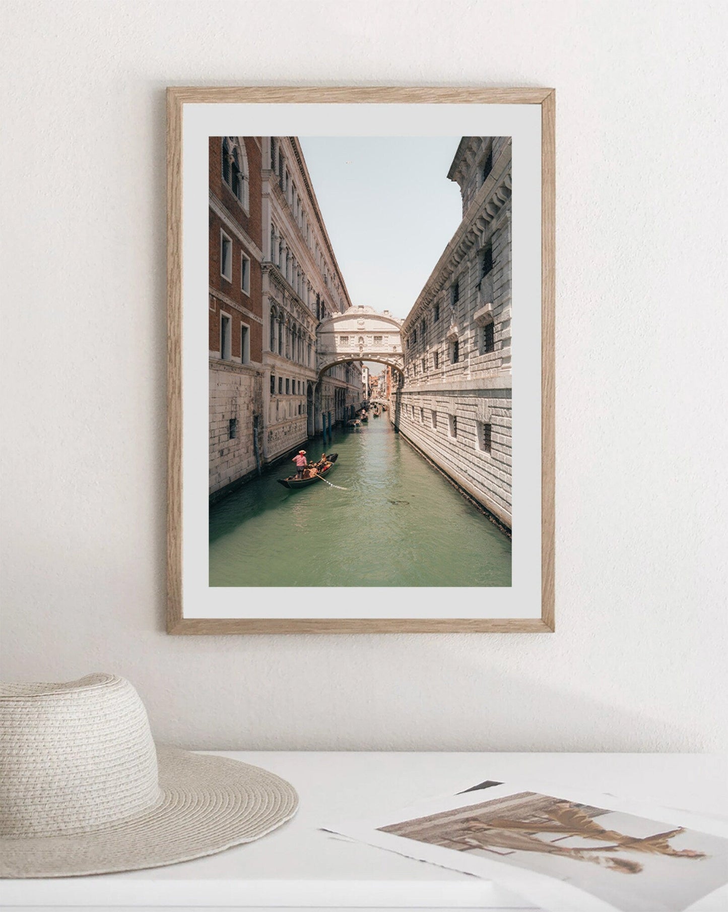 Framed Bridge of Sighs photo with a gondola scene, hanging on a white wall above a hat and books.