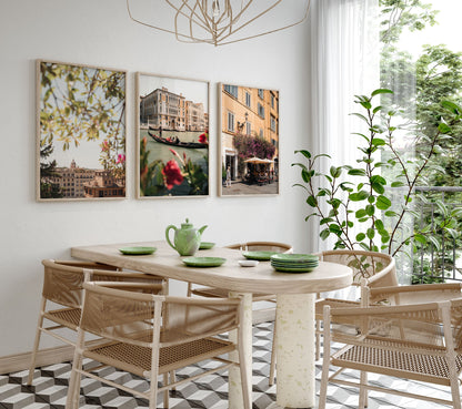 Modern dining room with a wooden table, four chairs, and Bright Day in Italy - Set of 3 wall art by a window.