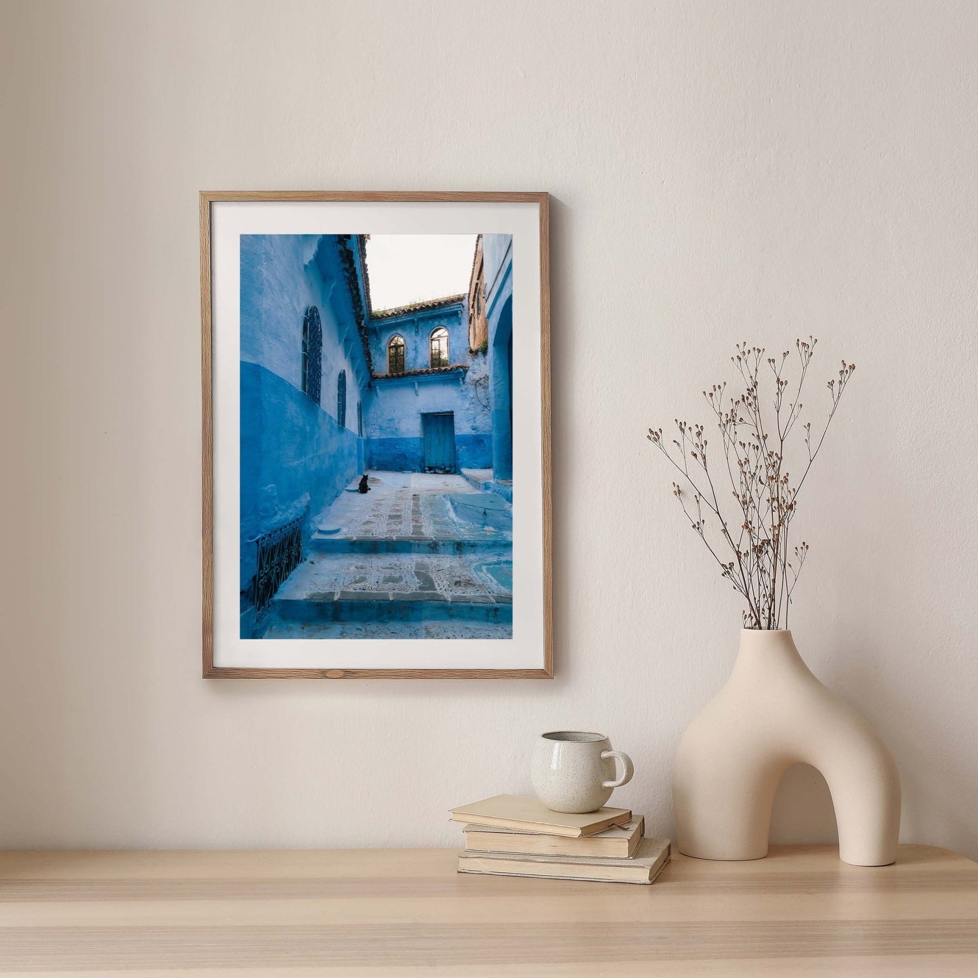 The Chefchaouen Cat framed photo hangs on a beige wall beside a vase and books.