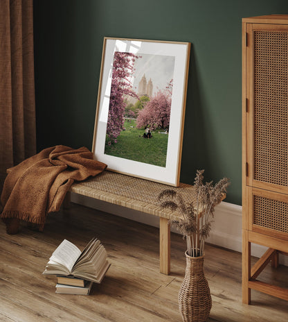 Framed Cherry Blossom I photo on woven bench, blanket, open book on floor, vase with dried flowers, green wall.