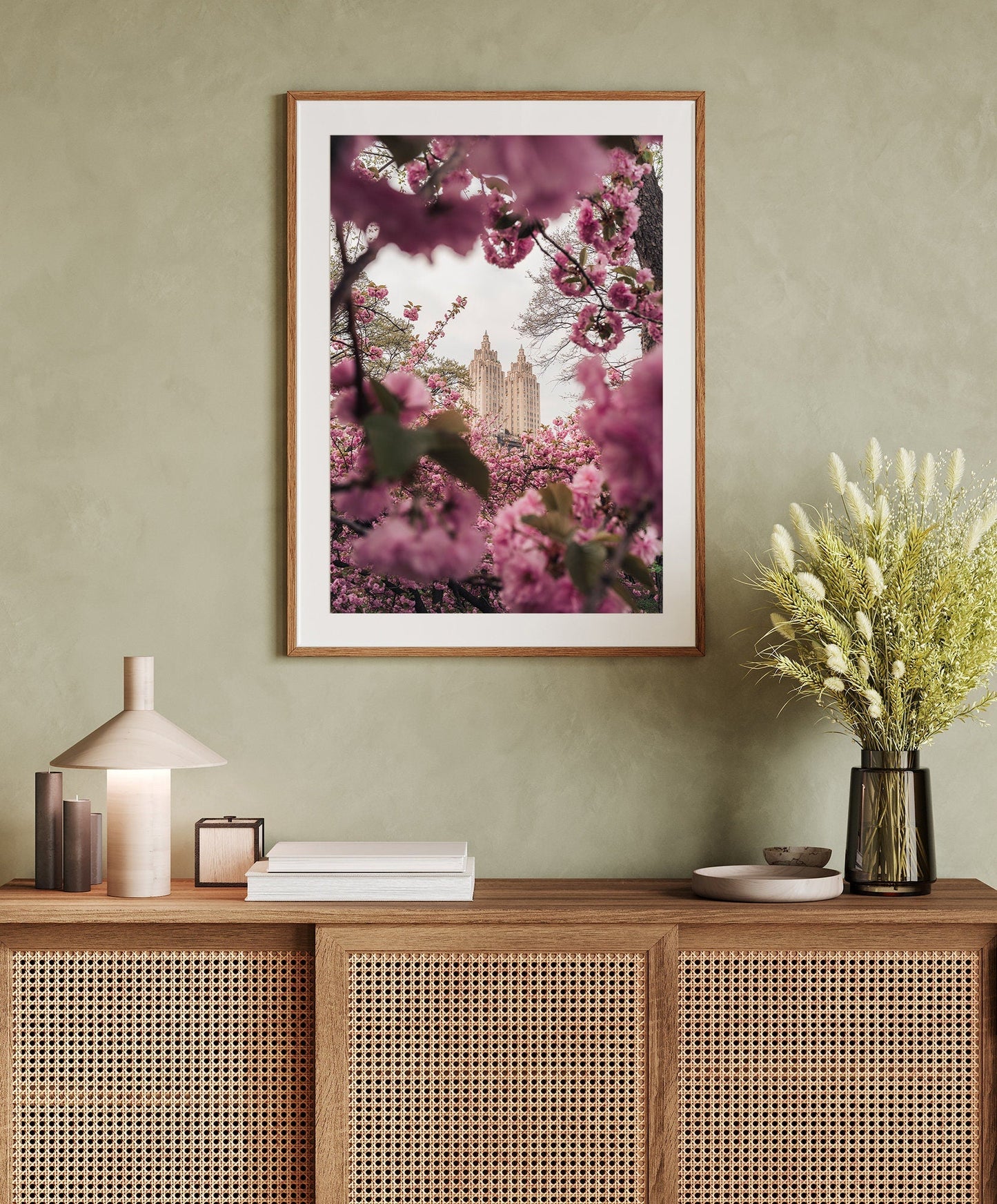 Cherry Blossom II on a green wall above a wooden sideboard with books, a lamp, and flowers.