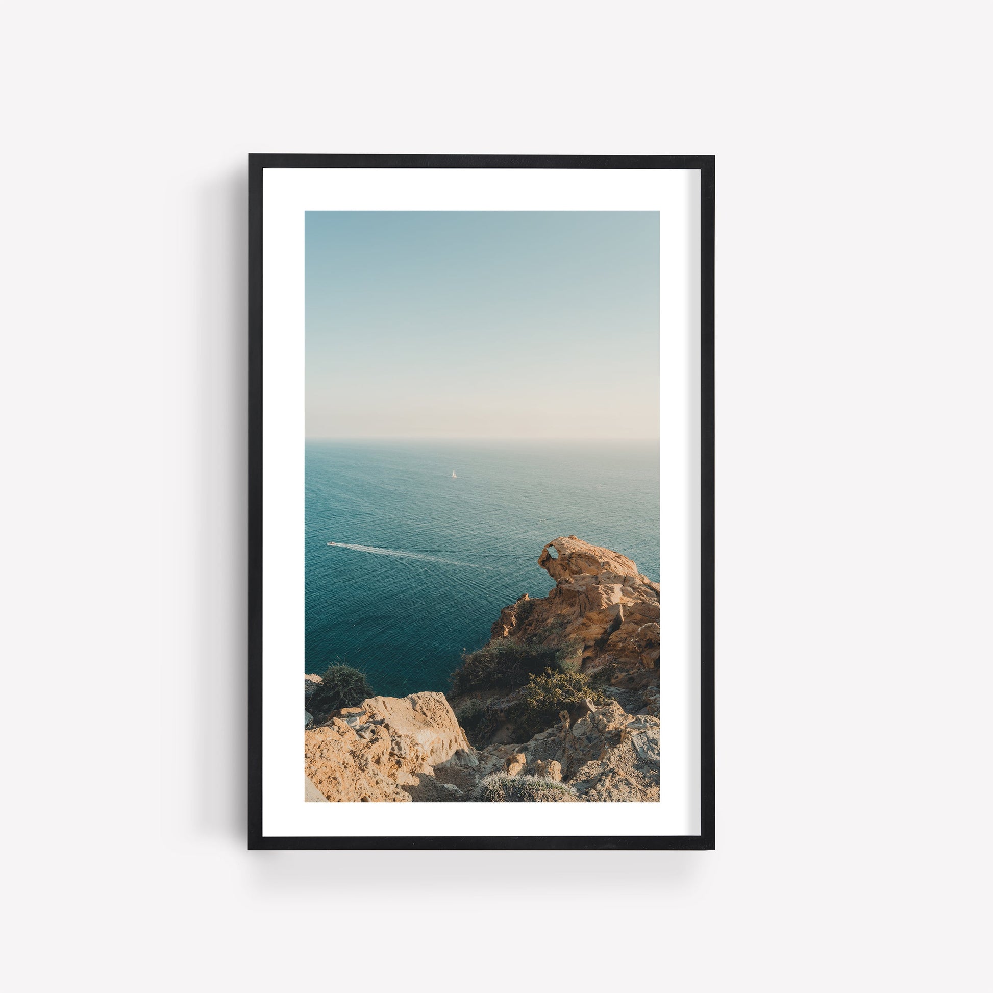 Cliffside Paradise framed photo: rocky cliff above calm blue ocean beneath a clear sky, displayed on a white wall.