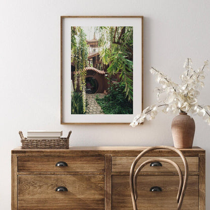 The Earthy Enlightenment wooden dresser has a framed photo, dried flowers in a vase, and a wicker basket on it, with a chair in front.