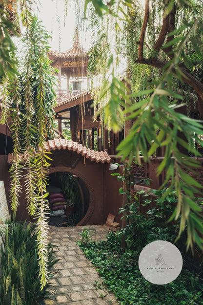 A lush garden path leads to Earthy Enlightenment, a wooden structure with greenery and red-tiled roofs.