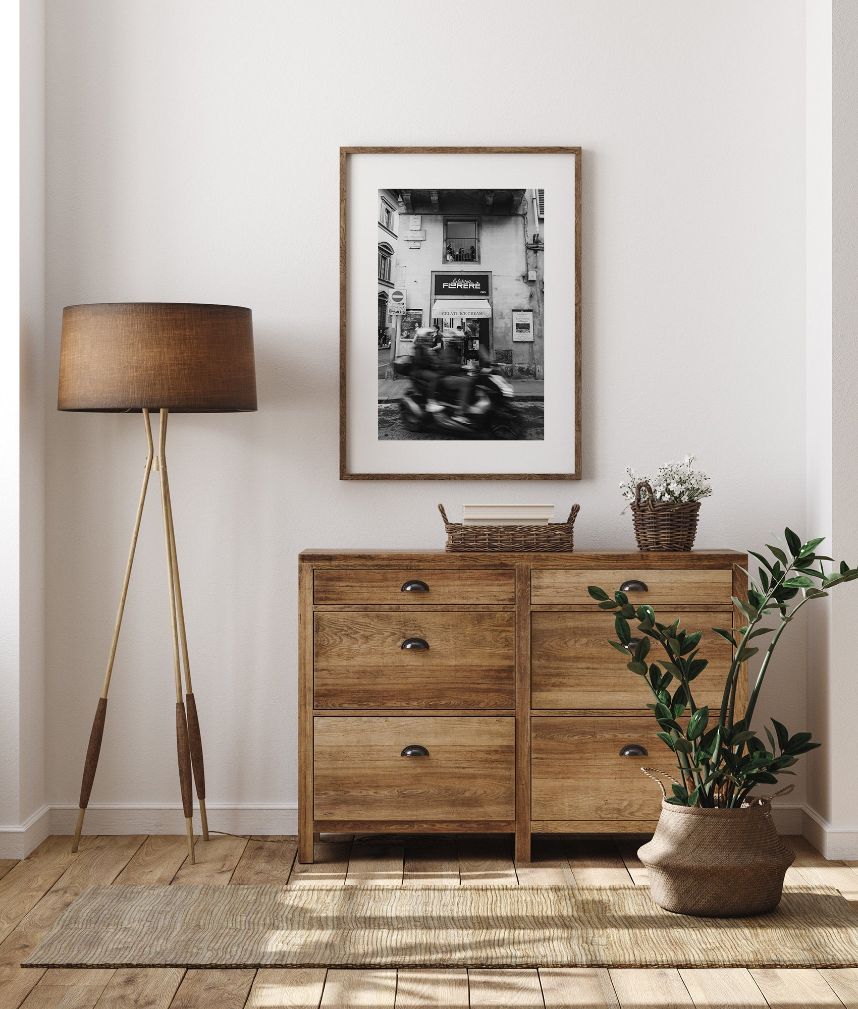 Cozy room with a wooden dresser, Florence Motorcycle art, tripod lamp, potted plant, and woven baskets on wood floor.