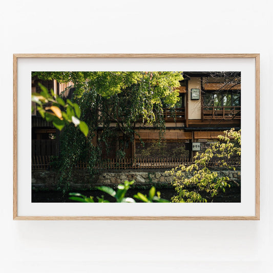 The Gion Greenery product is a framed photograph showcasing a traditional wooden building enveloped by lush greenery.