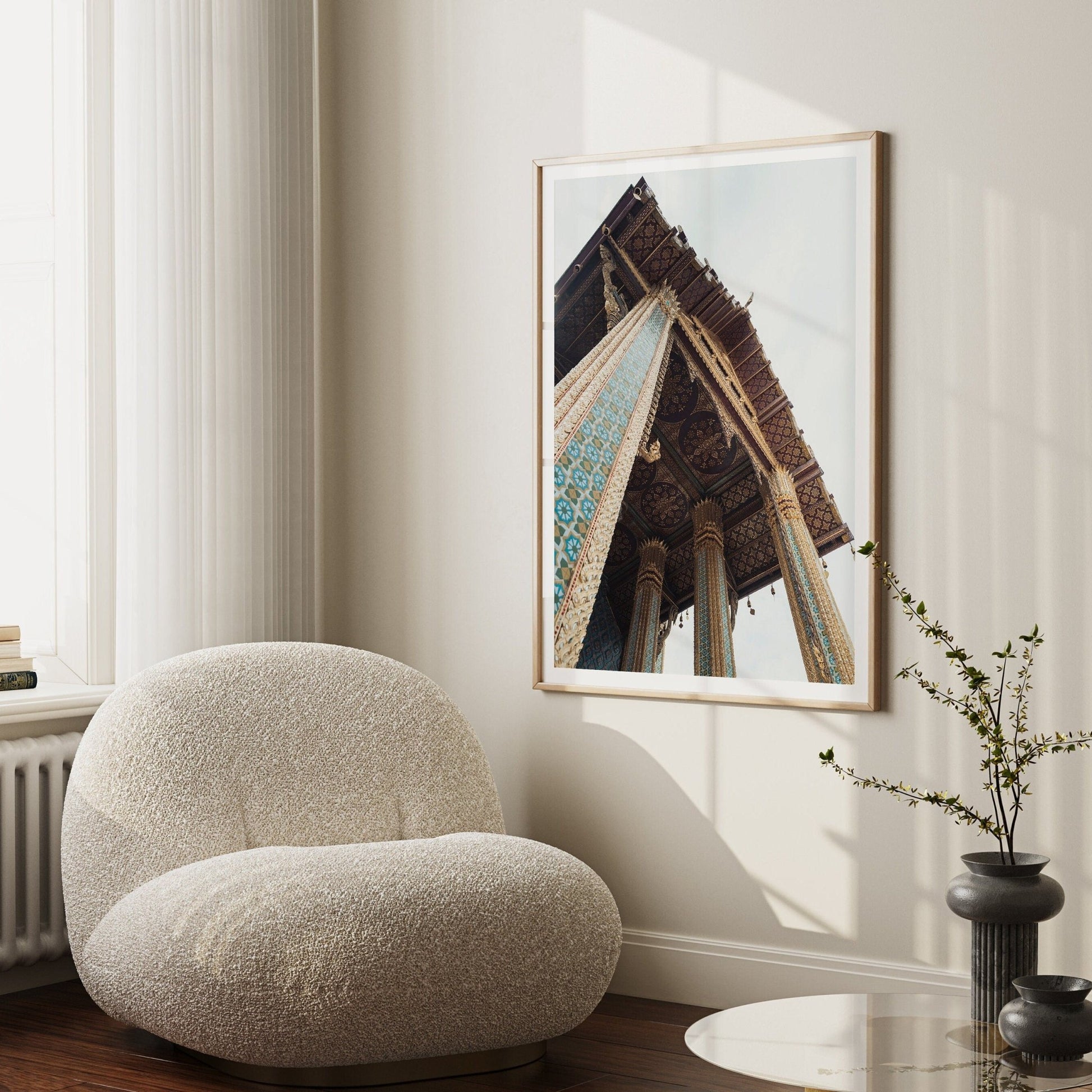 The Grand Palace II chair sits beside a table with a plant, beneath an architectural photo on a beige wall, basking in filtered sunlight.