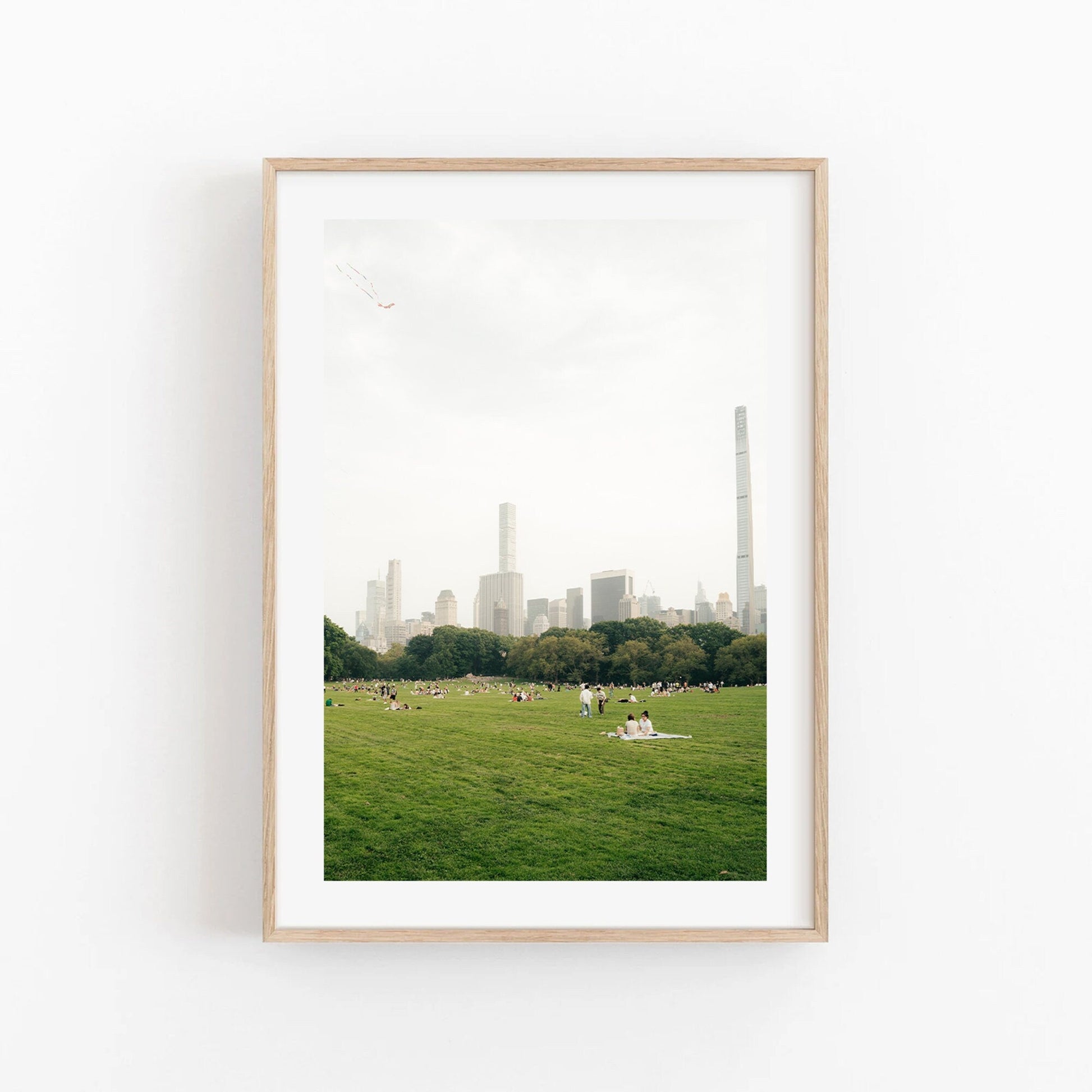 Framed photo titled Great Lawn II: city skyline with people on a grassy field in the foreground.