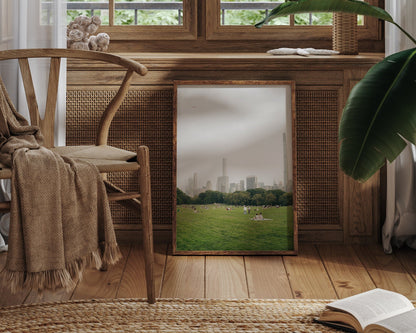 Great Lawn II mirror reflects a park scene with skyscrapers, next to a cozy chair, blanket, and book in a rustic room.