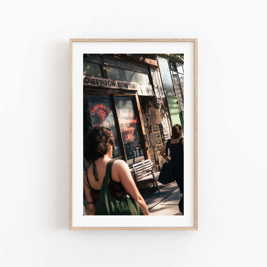 Two people walk past the Overthrow Boxing Club storefront with posters and graffiti, framed in wood on a white wall.